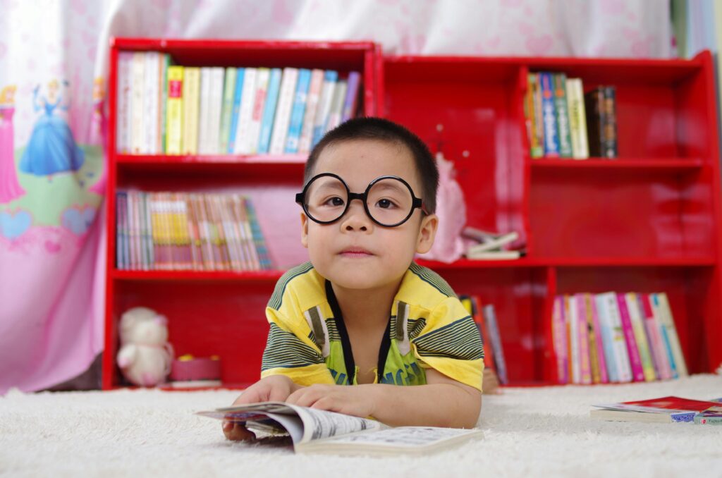 pexels-photo-261895-261895 A cute child in glasses reading a book, surrounded by colorful shelves. Perfect shot of childhood learning indoors.